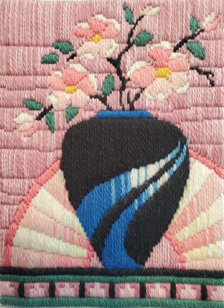 A longstitch needlepoint piece consisting of a black and blue art deco vase, with pink flowers, pink fan, and pink background