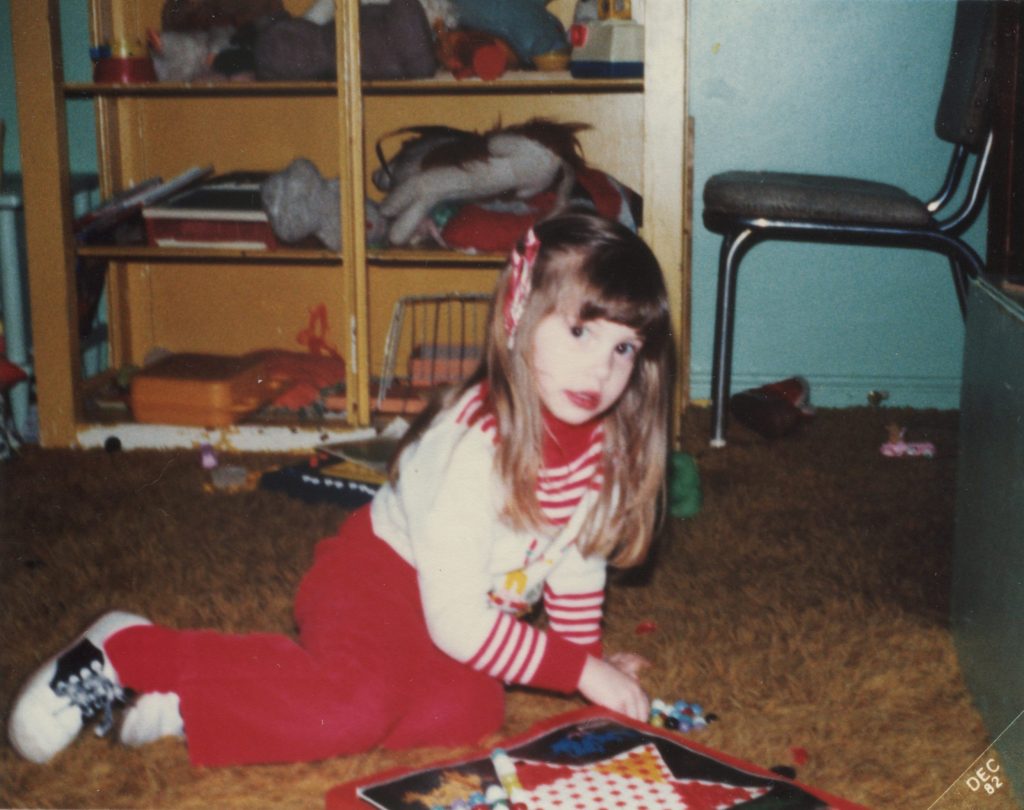 Lindsay at age 3, sitting on the shag carpet floor with a Chinese checkers board. She is wearing red pants, a white and red sweater and saddle shoes.